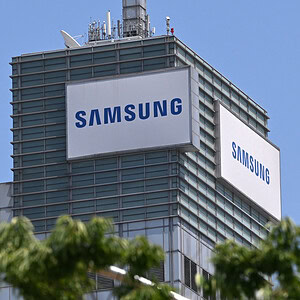 Modern kantoorgebouw van Samsung met grote logo's op de gevel, gelegen in een stedelijke omgeving onder een heldere blauwe hemel, omgeven door groene bomen.