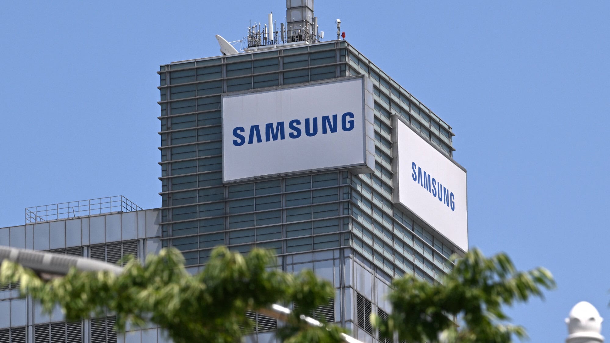 Modern kantoorgebouw van Samsung met grote logo's op de gevel, gelegen in een stedelijke omgeving onder een heldere blauwe hemel, omgeven door groene bomen.